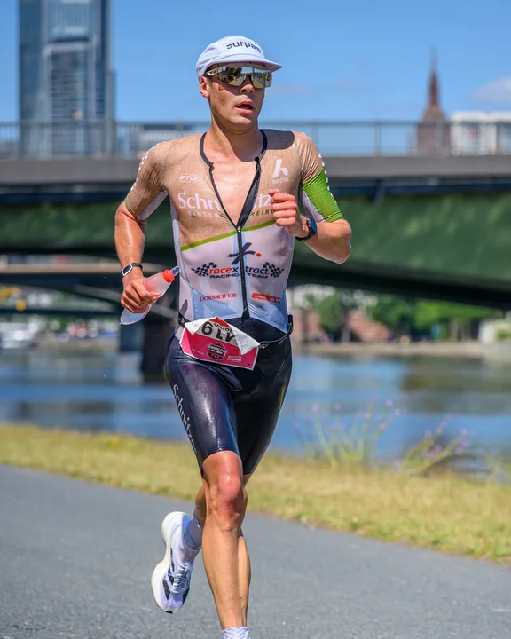 Der Triathlet Jonas Hoffmann l&auml;uft mit der Startnummer 6 auf einem Asphaltweg entlang eines Flusses. Er tr&auml;gt einen gesponserten Trisuit mit den Logos &bdquo;Schnitzer" und &bdquo;racex Racing Team", dunkle Laufshorts sowie wei&szlig;e Socken und Laufschuhe. In der Hand h&auml;lt er eine Trinkflasche, das Gesicht ist durch Sonnenbrille und wei&szlig;e Laufm&uuml;tze gesch&uuml;tzt. Im Hintergrund sind eine Br&uuml;cke, ein Hochhaus und ein Kirchturm bei strahlendem Sonnenschein zu erkennen.