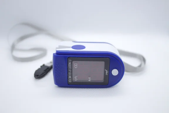 Ein blau-weißes Fingerpulsoximeter liegt auf einer hellen Fläche. Das Gerät ist ausgeschaltet, das Display zeigt keine Messwerte. An der Seite ist ein grauer Umhängeband befestigt. Auf dem Gehäuse sind die Bezeichnungen „Fingertip Oximeter" sowie die Messgrößen SpO₂ und BPM zu lesen.