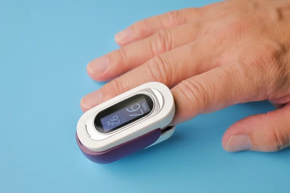 Eine Hand liegt auf hellblauem Untergrund. Am Zeigefinger ist ein weißes Pulsoximeter mit lila Unterseite befestigt. Das Display zeigt eine Sauerstoffsättigung von 97 % sowie einen Puls von 92 Schlägen pro Minute an.