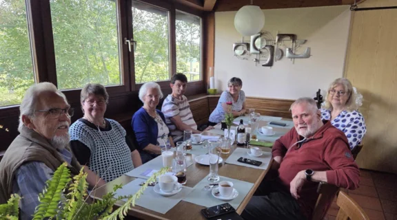 Sieben Mitglieder der Regionalgruppe Franken sitzen zusammen an einer gedeckten Tafel. Drei von ihnen tragen ein Sauerstoffger&auml;t. Auf dem Tisch stehen Kaffeetassen, Getr&auml;nke und eine kleine Blumenvase. Im Hintergrund sind gro&szlig;e Fenster mit Blick ins Gr&uuml;ne zu sehen.