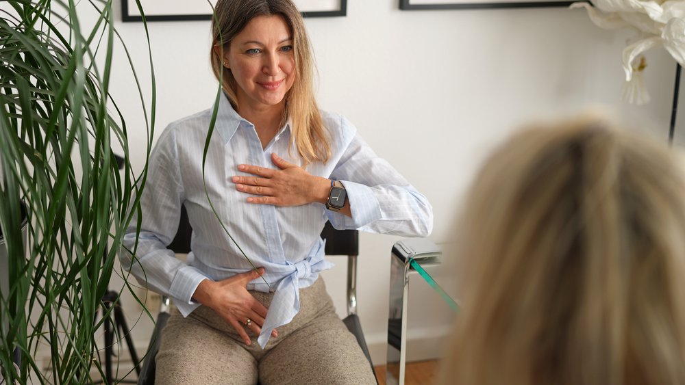 Atemübung Eine lächelnde Therapeutin sitzt einer Patientin gegenüber und demonstriert eine Atemübung: eine Hand liegt auf der Brust, die andere auf dem Bauch. Der Raum ist hell und freundlich eingerichtet.
