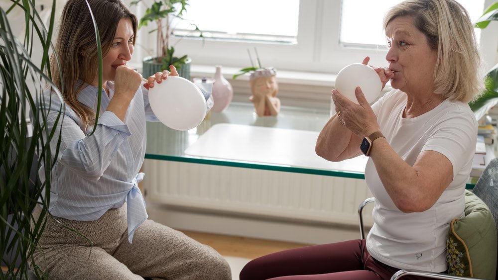 Therapeutin und Patientin &uuml;ben gemeinsam eine Atem&uuml;bung mit wei&szlig;en Luftballons in einem freundlichen Therapieraum.