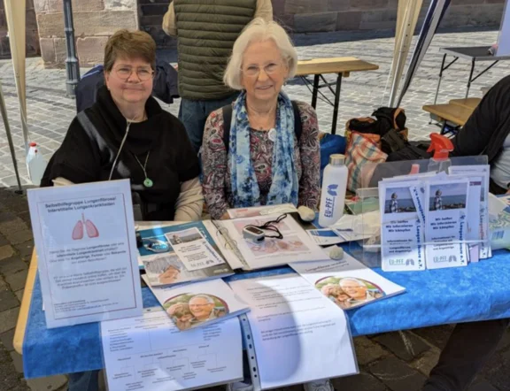 Zwei Frauen der Regionalgruppe Franken informieren mit einem Marktstand in N&uuml;rnberg &uuml;ber die Lungenfibrose. Auf dem Tisch vor ihnen iegen Flyer und Brosch&uuml;ren aus.