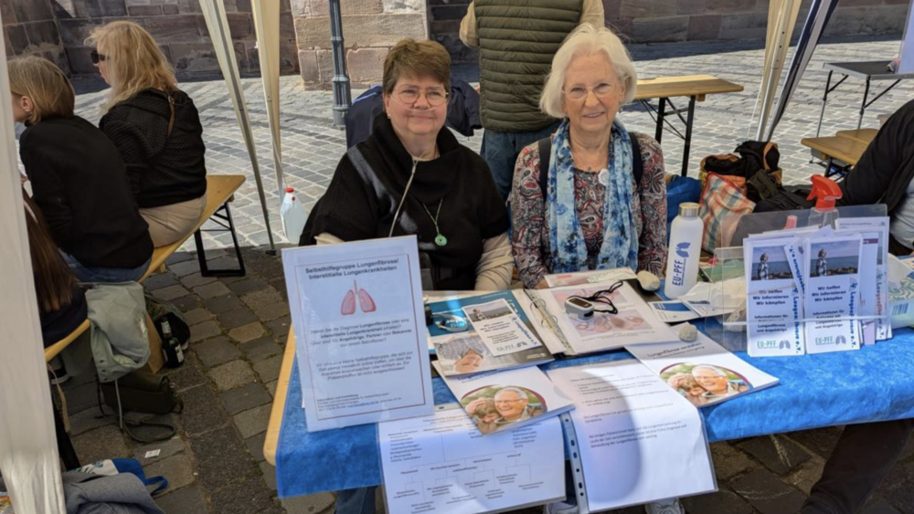 Zwei Frauen der Regionalgruppe Franken informieren mit einem Marktstand in N&uuml;rnberg &uuml;ber die Lungenfibrose. Auf dem Tisch vor ihnen iegen Flyer und Brosch&uuml;ren aus.