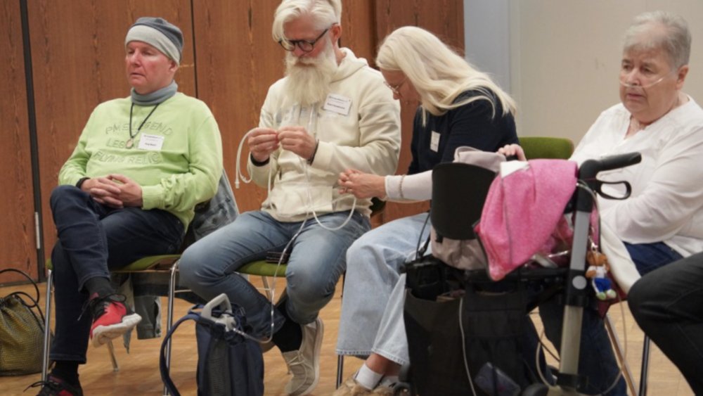 Eine Veranstaltung des Lungenfibrose e.V. F&uuml;nf Personen sitzen in einem Veranstaltungsraum. Eine Teilnehmerin mit Sauerstoffger&auml;t und Rollator ist dabei. Zwei Teilnehmende machen sich mit einem mobilen Sauerstoffger&auml;t mit Schlauch vertraut. Das Ger&auml;t befindet sich in einem kleinen Rucksack.