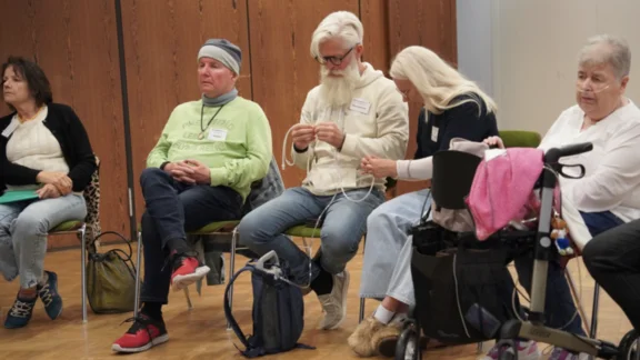 Mobiles Sauerstoffgerät Eine Veranstaltung des Lungenfibrose e.V. Fünf Personen sitzen in einem Veranstaltungsraum. Eine Teilnehmerin mit Sauerstoffgerät und Rollator ist dabei. Zwei Teilnehmende machen sich mit einem mobilen Sauerstoffgerät mit Schlauch vertraut. Das Gerät befindet sich in einem kleinen Rucksack.